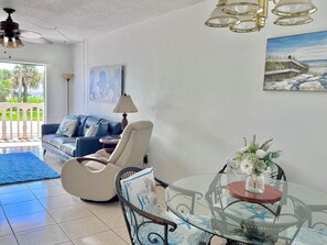 A living room with a glass table, a blue sofa, a white armchair, and a painting on the wall. - Oceanview Across From The Beach (Ormond by the sea)