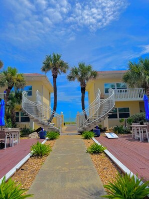 A pathway leading to a building with a spiral staircase, surrounded by palm trees and outdoor seating. - Oceanview Across From The Beach (Ormond by the sea)
