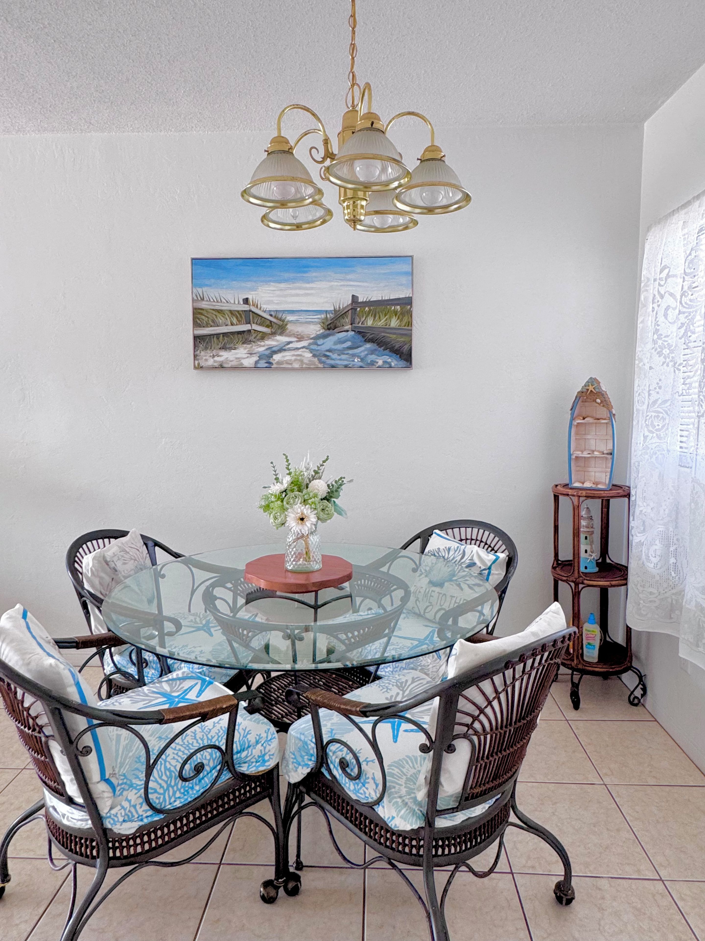 Un coin repas avec une table en verre entourée de chaises en osier, un tableau représentant une scène de plage et une lanterne décorative.