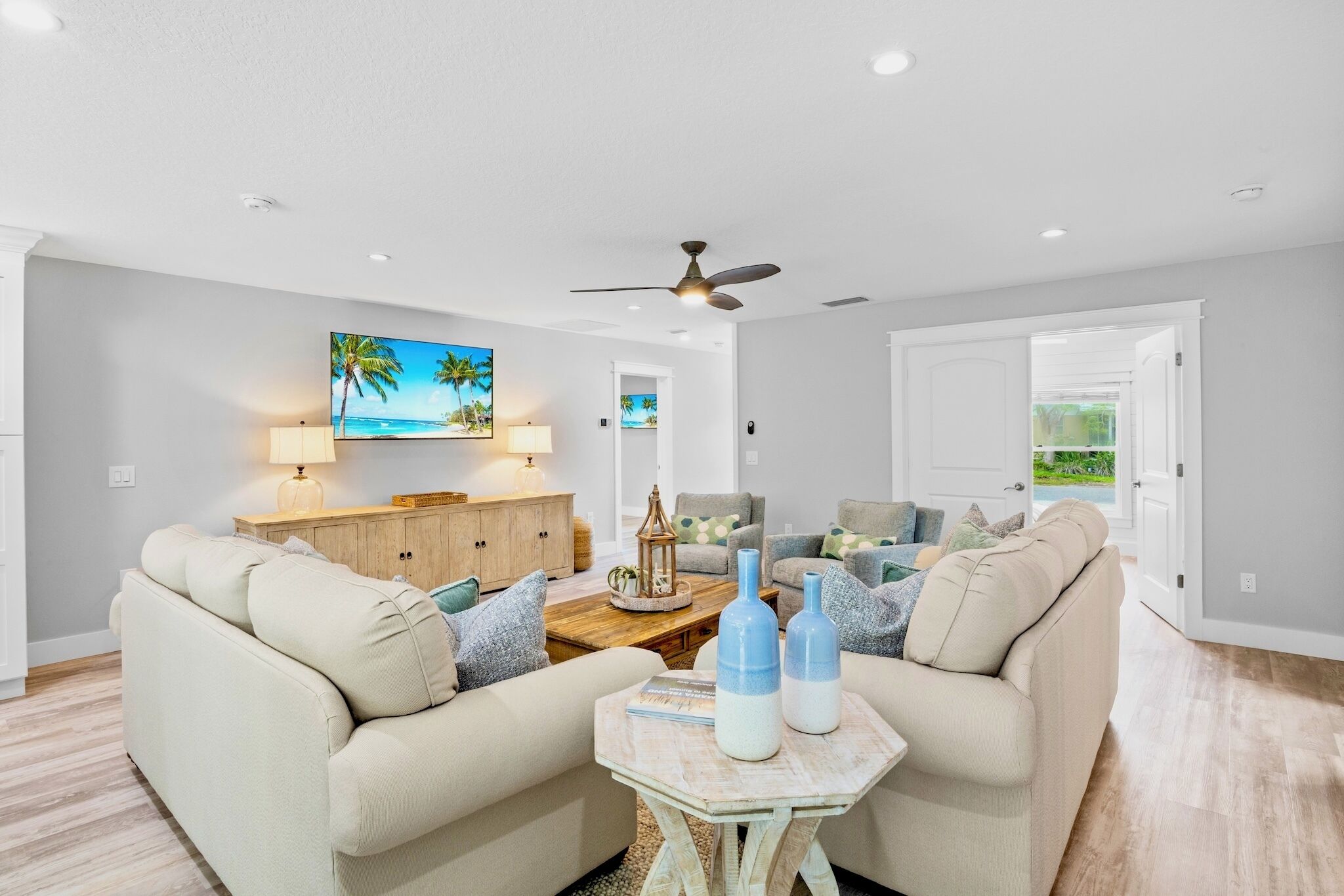 A living room with a beige sofa, a coffee table with decorative bottles, a TV mounted on the wall, and a ceiling fan.