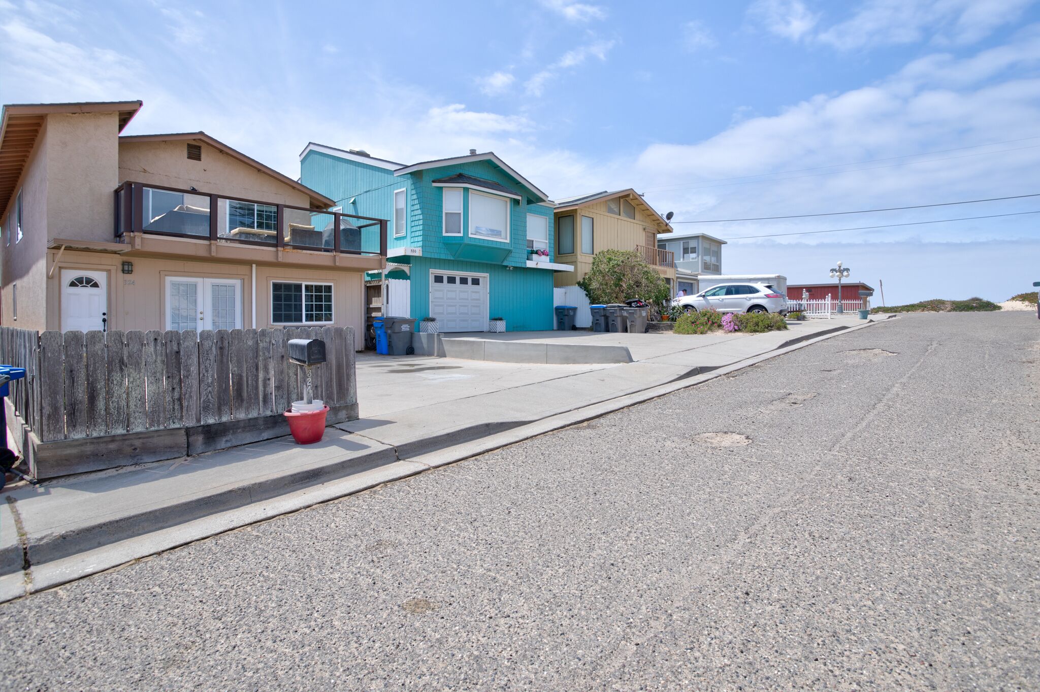Una calle residencial con casas, un coche blanco y una playa al fondo.
