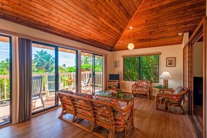 A spacious living area with a wooden ceiling, large sliding glass doors leading to a balcony, wicker furniture, and a television.