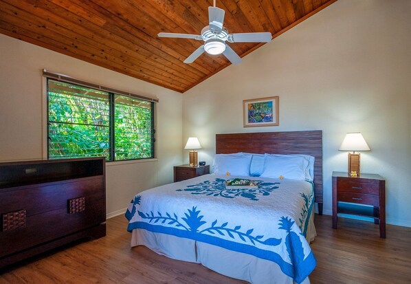 A bedroom with a wooden ceiling, a bed with a blue floral bedspread, two bedside lamps, and a window with a view of greenery.