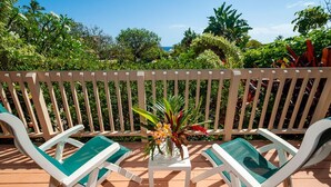 A covered outdoor seating area with two white lounge chairs and a small table, surrounded by a wooden fence and lush greenery.