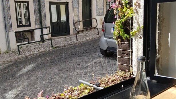 En utsikt från ett fönster mot en kullerstensgata med byggnader, en parkerad bil och en flaska på ett bord.