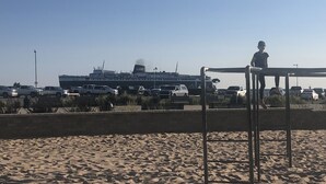 Una playa arenosa con una estructura metálica, un barco grande al fondo y coches aparcados.