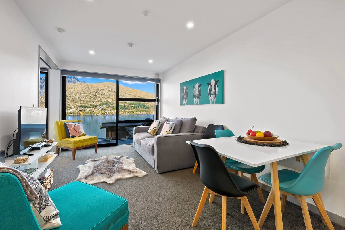 A modern living room with a sofa, a yellow armchair, a dining table with chairs, and a large window offering a view of a mountainous landscape.