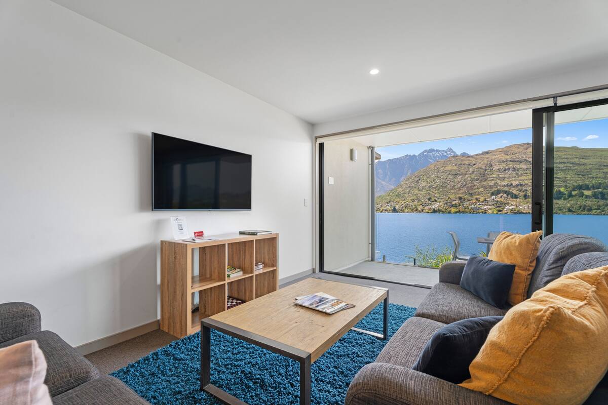 A modern living room with a large flat-screen TV, a wooden bookshelf, a blue rug, a gray sofa with yellow cushions, and a view of a lake and mountains through a glass door.