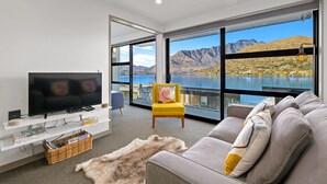 Una sala de estar moderna con un televisor de pantalla plana grande, un sofá, una silla amarilla y vistas a montañas y un lago a través de amplias ventanas.