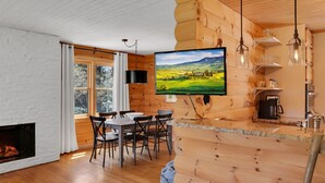 A cozy cabin interior with a dining area, a fireplace, a TV mounted on a wooden wall, and a kitchen area with a coffee machine. - The Kind of Place You Stop Rushing the Moment You Arrive | Bungalow on Crowe (Highlands)