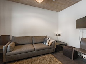 Een moderne woonkamer met een bank, een bijzettafeltje, een lamp en een aan de muur bevestigde televisie.