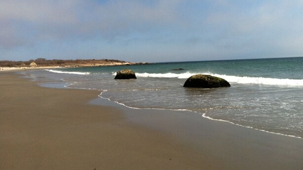 Una playa con olas tranquilas, orilla de arena y algunas rocas.