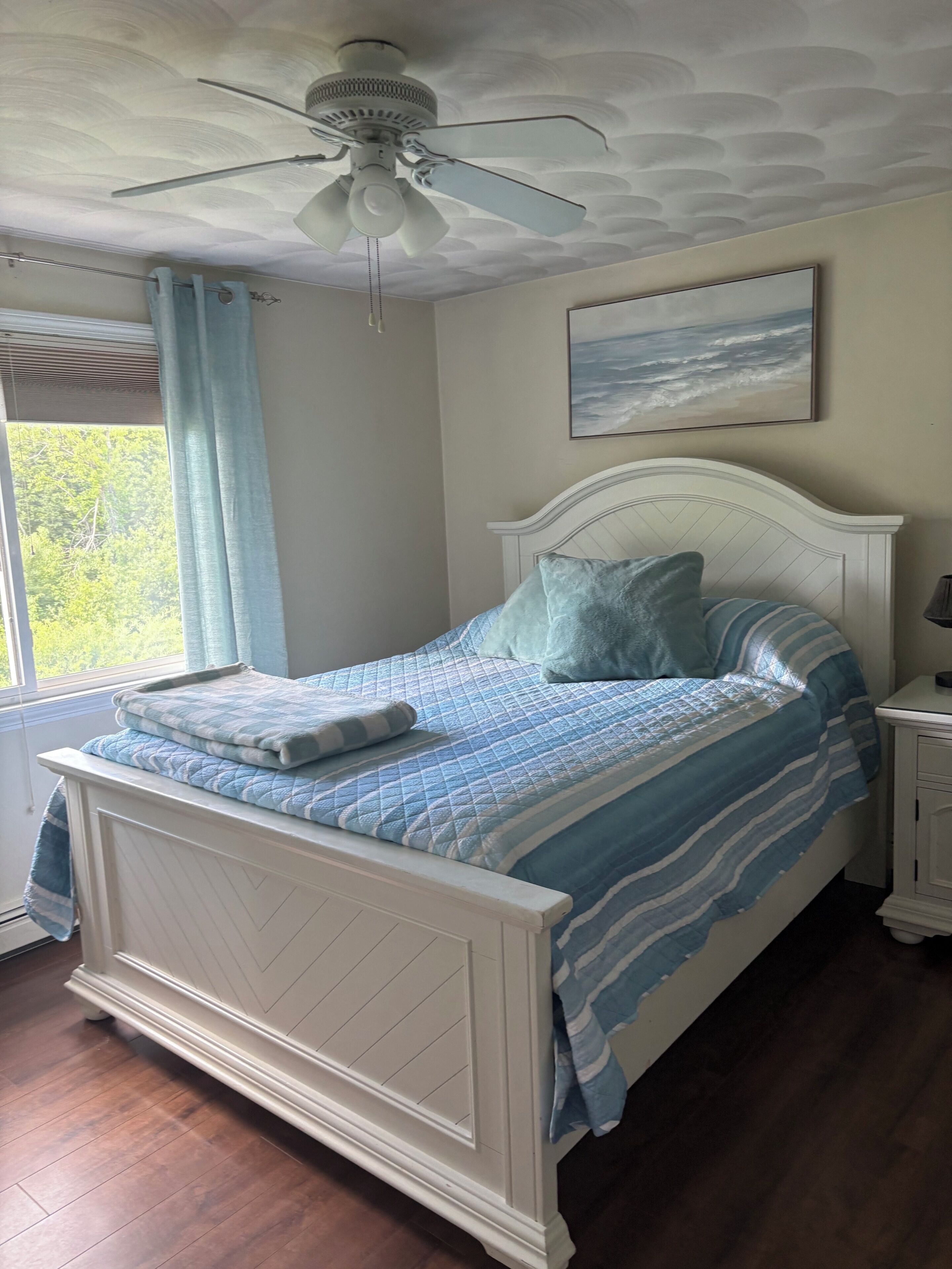 A bedroom with a bed, a ceiling fan, a window with curtains, a nightstand, and a painting on the wall.