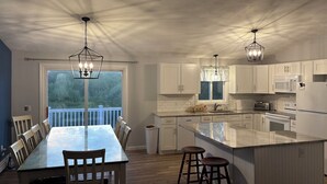 Una cocina moderna con armarios blancos, una isla grande con taburetes, una mesa de comedor con sillas y lámparas colgantes.