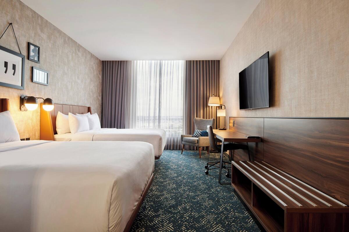 Ein Hotelzimmer mit zwei Betten, einem Schreibtisch, einem Stuhl, einem Fernseher und einem Fenster mit Vorhängen.