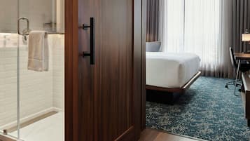 Ein Hotelzimmer mit Holzschrank, Duschkabine aus Glas, Bett mit blau gemustertem Teppich und Schreibtisch mit Stuhl.