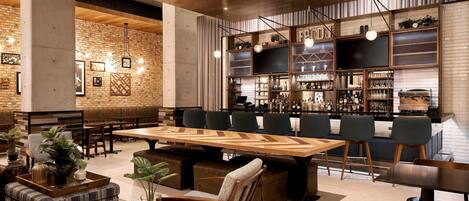 Un moderno espacio de bar con una mesa de madera larga, taburetes y un fondo de pared de ladrillo.