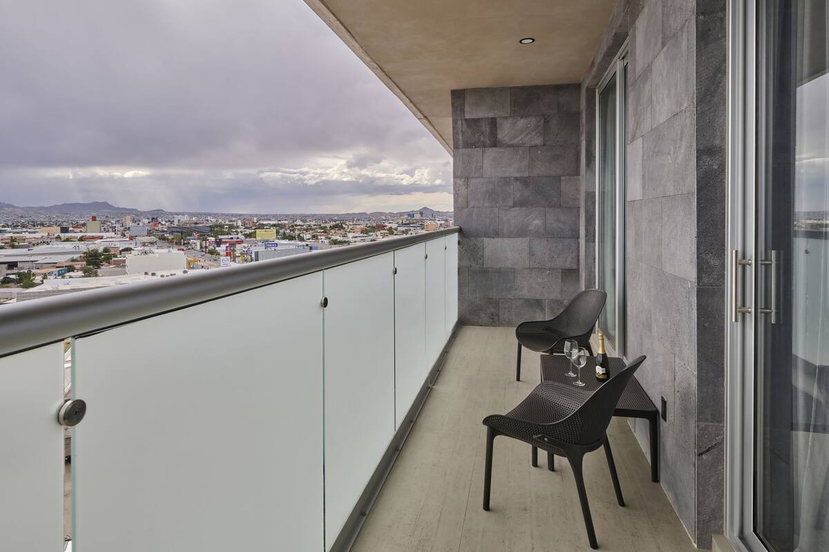 Ein Balkon mit zwei Stühlen und einem Tisch, Blick auf die Stadt und ein großes Fenster.