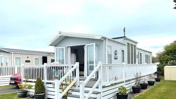 A mobile home with a porch and a small deck, surrounded by potted plants and a parked car. - Luxury 3 bed mobile home on the sea (Southampton)