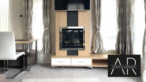 A modern living room with a flat-screen TV mounted on a wooden wall, a fireplace below, and a seating area with a sofa and a chair. - Luxury 3 bed mobile home on the sea (Southampton)