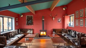 A cozy interior with red walls, a central wood-burning stove, and cowhide upholstered seating. - 1 Bed Shepherds Hut - Sleeps 2 -Wood Fired Hot Tub (Charlton musgrove)