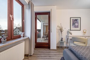 Une chambre d’hôtel comprenant un lit, un bureau, une chaise, une fenêtre avec des rideaux, un radiateur et un tableau encadré au mur.