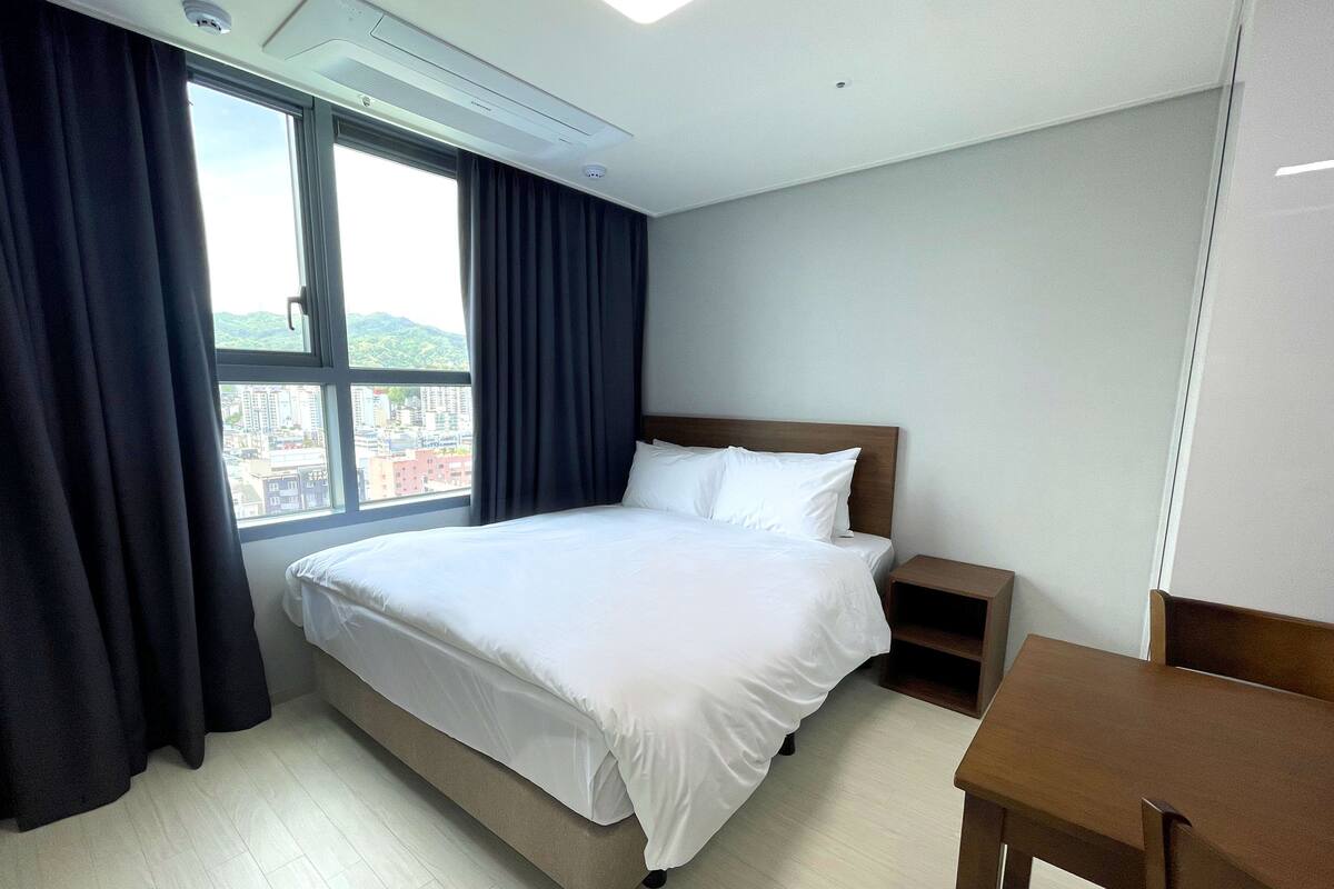 Une chambre d’hôtel avec un grand lit, une table de chevet en bois, une petite table et une vue sur le paysage urbain par une fenêtre ornée de rideaux foncés.