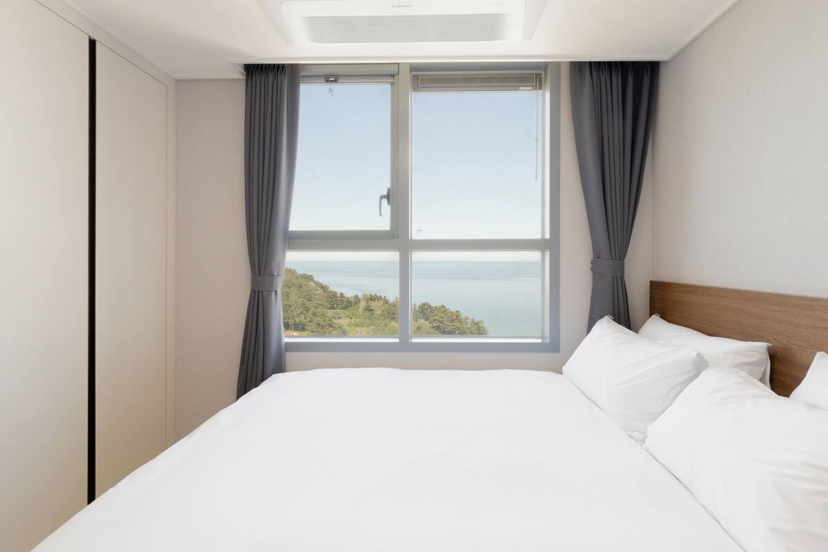 Une chambre dotée d’une grande fenêtre, d’un lit avec du linge de lit blanc et offrant une vue sur un paysage naturel.