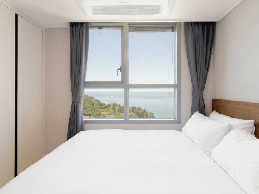 A bedroom with a large window, a bed with white linens, and a view of a natural landscape.