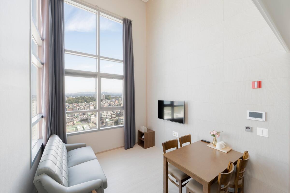Un salon moderne doté d’une grande fenêtre, d’un canapé, d’une table à manger avec des chaises, d’un téléviseur à écran plat et d’une petite table d’appoint.
