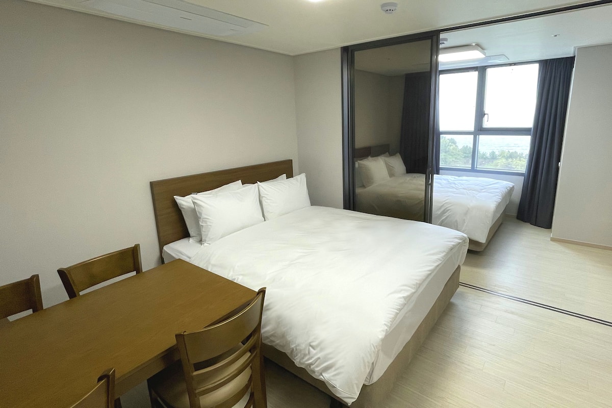 Une chambre d’hôtel avec un grand lit, une table en bois et une chaise. Une porte coulissante donne sur un balcon offrant une vue sur l’extérieur.
