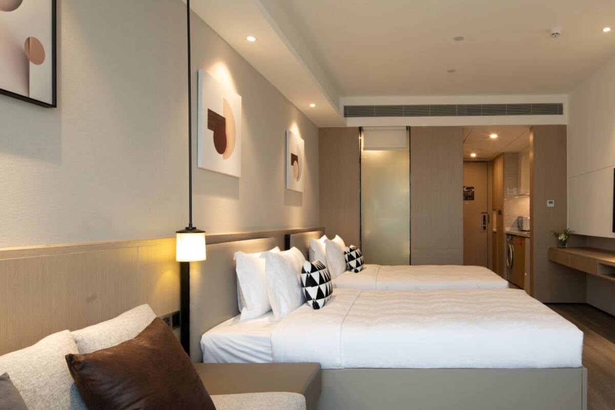 A modern hotel room with two beds, a sofa, and a bathroom visible through a sliding door.