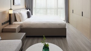 Una habitación de hotel moderna con una cama grande, una mesita de noche, una lámpara, un pequeño sofá, una mesa de centro con una planta y un ventanal con cortinas.