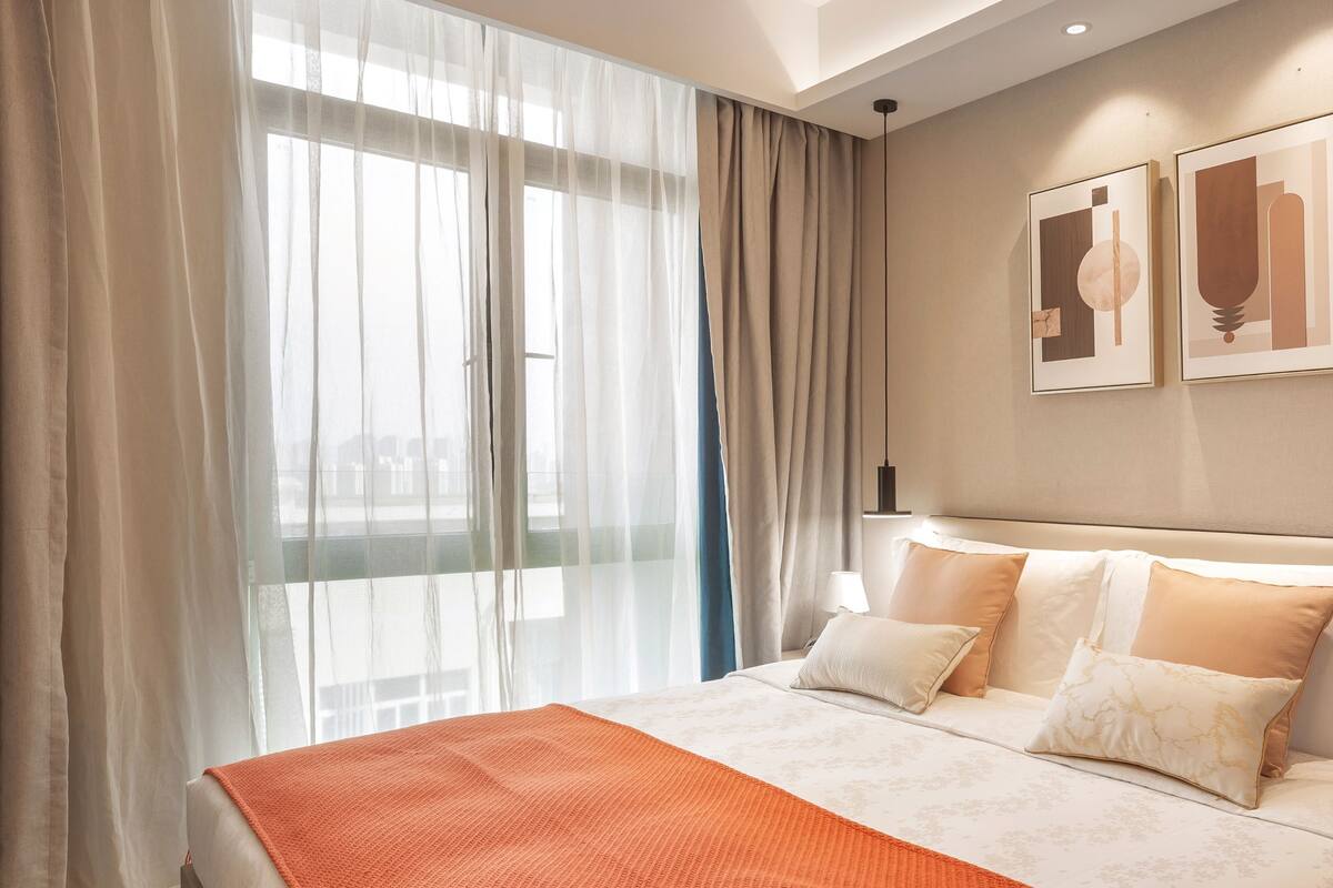 A hotel room with a large bed, a view through sheer curtains, and two framed artworks on the wall.