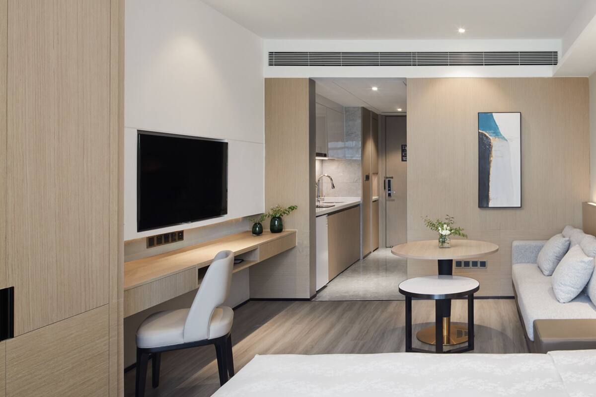 A modern hotel room with a large bed, a desk with a flat-screen TV, a seating area with a sofa and armchairs, and a small round table.