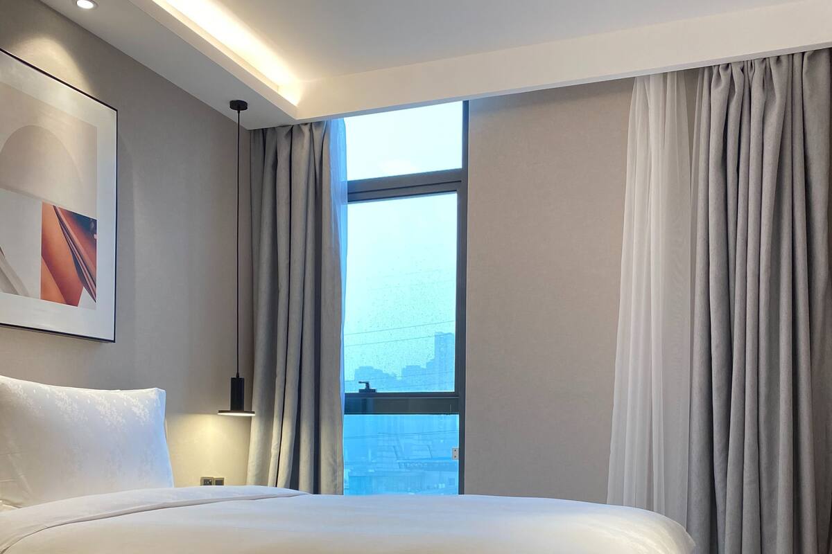A modern hotel room with a large bed, a bedside table, a lamp, and a window with curtains.
