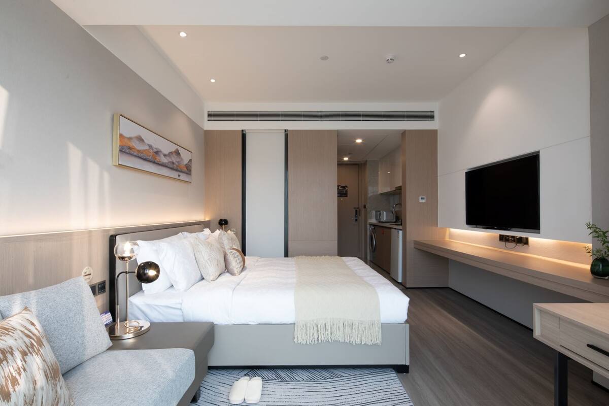 A modern hotel room with a bed, a bedside table, a TV, a sofa, and a kitchen area.