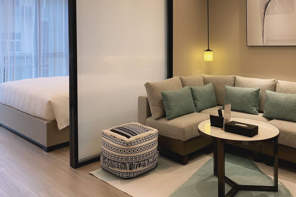 A modern hotel room with a bed, a sofa, a small ottoman, a round table, and a large window with sheer curtains.