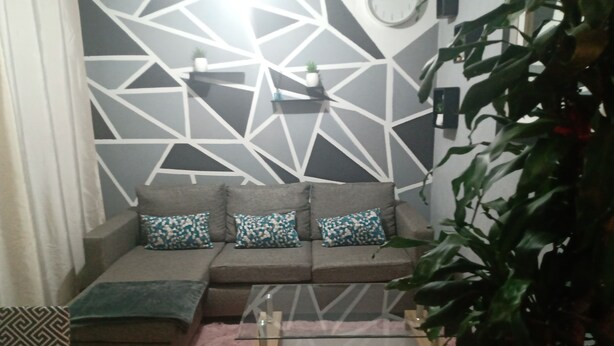 Ein Wohnzimmer mit grauem Sofa, gemusterten Kissen, einem Glastisch und einem geometrischen Wanddesign.