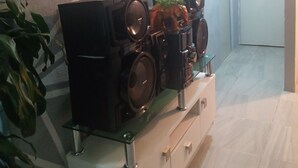 Ein Zimmer mit moderner Einrichtung, das eine Wand mit geometrischen Mustern, einen Konsolentisch mit Glasplatte und Lautsprechern sowie eine Tür zu einem weiteren Raum zeigt.