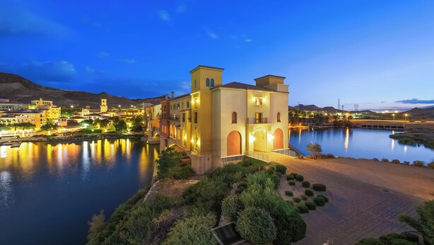 A historic building with arched windows and a tower, surrounded by a calm lake and well-maintained gardens.