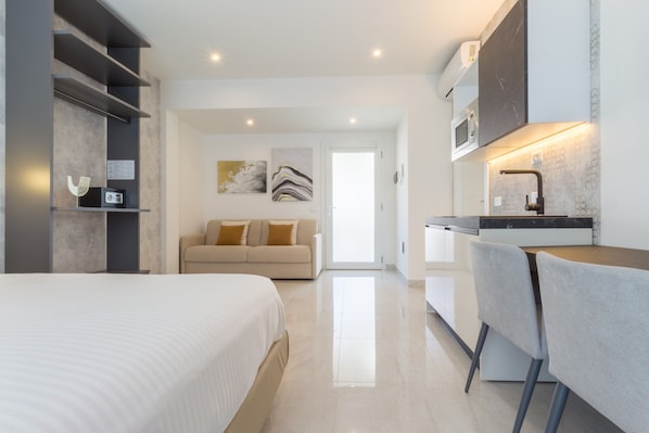 A modern hotel room with a bed, a sofa, a kitchenette, and a bathroom.