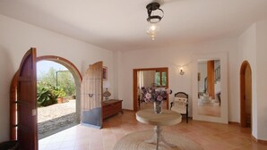 A spacious hallway with a tiled floor, a wooden bench, a round table with a vase, and a view of the garden through an open archway. - Chalet "Sa Corbaia Nova" with Mountain View, Pool, Wi-Fi, Terraces & Garden (Artà)