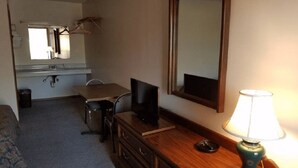 Une chambre d’hôtel de taille réduite, comprenant un lit simple, un petit bureau avec une télévision, un miroir, une lampe et une fenêtre donnant sur le ciel.