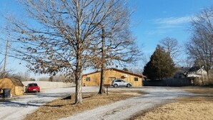 Un cadre rural avec une maison en bois, une allée en gravier et une voiture rouge garée devant.
