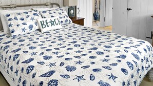 Un dormitorio con una cama con colcha náutica azul y blanca, una mesita de noche con lámpara, y una puerta que da a otra habitación.