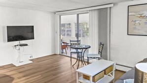 Un salón moderno con televisor de pantalla plana, suelo de madera, sofá blanco, mesa de comedor redonda con sillas y un balcón con vistas al exterior.