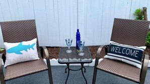 Dos sillas de mimbre con cojines, una mesa pequeña con un vaso y una botella azul, y una cerca blanca al fondo.