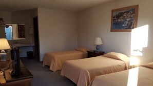 A room with two beds, a desk with a lamp, a painting on the wall, and a window providing natural light.