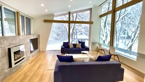 A modern living room with a fireplace, a dark-colored sofa set, and large windows offering a view of snow-covered trees. - Sanzan Chalet (Hakuba)
